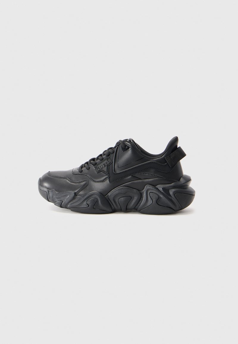 Svart chunky sneakers med bølgete såle, snøring og drageløft bak, vist mot en enkel hvit bakgrunn.