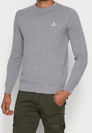 Uomo che indossa un maglione a maniche lunghe grigio chiaro con una piccola logo bianco e pantaloni cargo verdi, una mano in tasca.