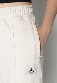 Närbild av en hand i fickan på ljusbeiga sweatpants med elastisk midja och en svart Jumpman Flight-logotyp. 