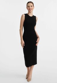 Czarna, prążkowana sukienka dzianinowa o średniej długości, z falbaniastym brzegiem u dołu i bez rękawów. Nosi dodatki w postaci beżowych szpilek.