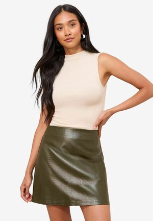 Vrouw met lang zwart haar in een mouwloos beige top en een groene leren rok met textuur, poserend met hand op heup tegen een witte achtergrond.