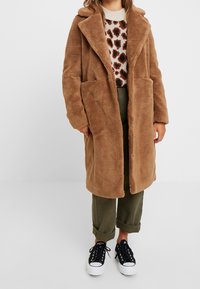Persona que lleva un abrigo de peluche marr�ón largo sobre un suéter de estampado de leopardo, pantalones oliva remangados y zapatillas en blanco y negro.