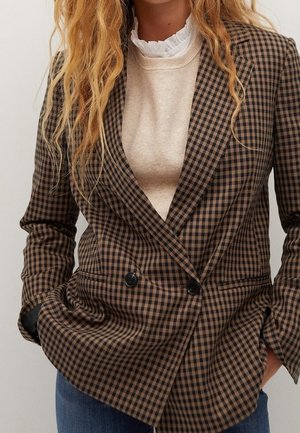 Woman wearing brown and black checkered double-breasted blazer over cream ruffled blouse and blue jeans, hands in pockets.