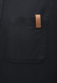 Haine albastru închis cu un buzunar pătrat vizibil. Prezintă o etichetă dreptunghiulară din piele maro cusută pe buzunar, cu inscripția „Pier One.”