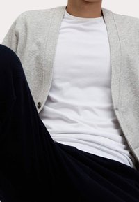 Cardigan grigio chiaro in tessuto a maglia, abbinato a una t-shirt bianca a collo rotondo, seduto con pantaloni navy a fantasia visibili.