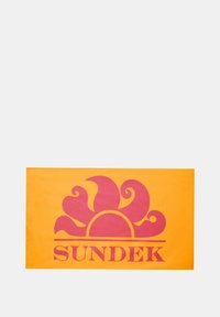 Asciugamano giallo con una grande grafica di un sole rosa e accenti a vortice, con la parola "SUNDEK" in lettere maiuscole in basso.