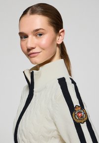 Vit kabelstickad zip-up tröja med svarta detaljer. Har hög krage och logotyp patch på vänster axel. Slät textur och figurnära design.