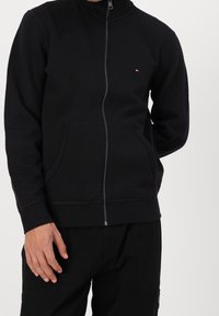Felpa nera con zip e colletto alto, dotata di due tasche frontali e branding discreto sul petto. Tessuto morbido e polsi a coste.