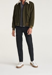 Giacca bomber verde con colletto in shearling, polo grigia, pantaloni cargo neri e scarpe da ginnastica bianche con suola marrone.
