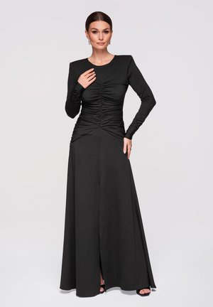 Kvinde iført lang sort rynket kjole med lange ærmer og en frontslids, stående selvsikkert foran en ensfarvet lys baggrund.