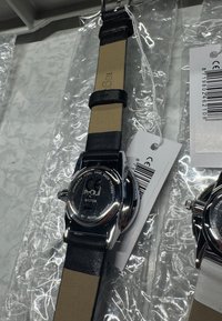 Silberne Armbanduhr aus Edelstahl mit schwarzem Lederarmband und beiger Unterseite. Durchsichtiger Rücken zeigt Markenlogo und Modellnummer. In Plastik versiegelt.