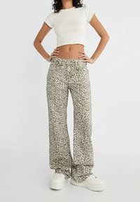 Kvinna som bär hög midja leopardmönstrade byxor, vitt kort t-shirt och vita plattformsskor, står med händerna på höfterna.