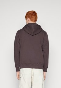 Sudadera marrón oscuro con capucha ajustable, puños acanalados y dobladillo. Hecha de tejido suave, combinada con pantalones cargo de color claro.