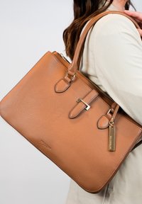 Bruine leren tote bag met een gestructureerd oppervlak, dubbele handvatten, zilveren accenten en een ritsluiting. Heeft een metalen label en een strak ontwerp.