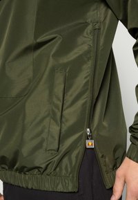 Žalias išorinis švarkas, pasižymintis glotnia, blizgia tekstūra, šoniniu užtrauktuko kišeniu, elastingais rankogaliais ir logotipu ant užtrauktuko traukimo.