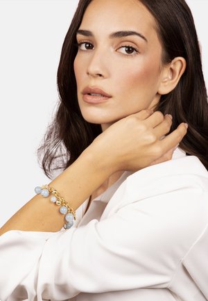 Gouden armband met blauwe kralen en metalen accenten, gedragen om een gebruinde pols. Witte kleding en een neutrale achtergrond. Glad oppervlak.