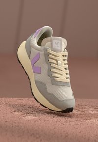 Sportssko med en beige mesh-överdel och grå och lavendel detaljer, krämfärgade snören och en strukturerad gummisula. Logotyp på tungan.