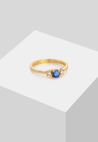 Anillo de oro con una piedra central azul flanqueada por dos pequeñas piedras transparentes, banda lisa, acabado pulido y diseño simple.