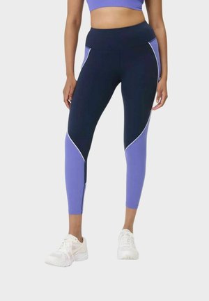 Vrouw draagt hooggetailleerde navy en paarse color-block legging, gecombineerd met witte sportschoenen, staand tegen een effen achtergrond.