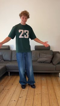 Žalias per didelis marškinėliai su baltu ir rožiniu numeriu 23, derinami su laisvais mėlynais džinsais ir juodais batais, stovintis ant medinių grindų šalia pilko sofos.