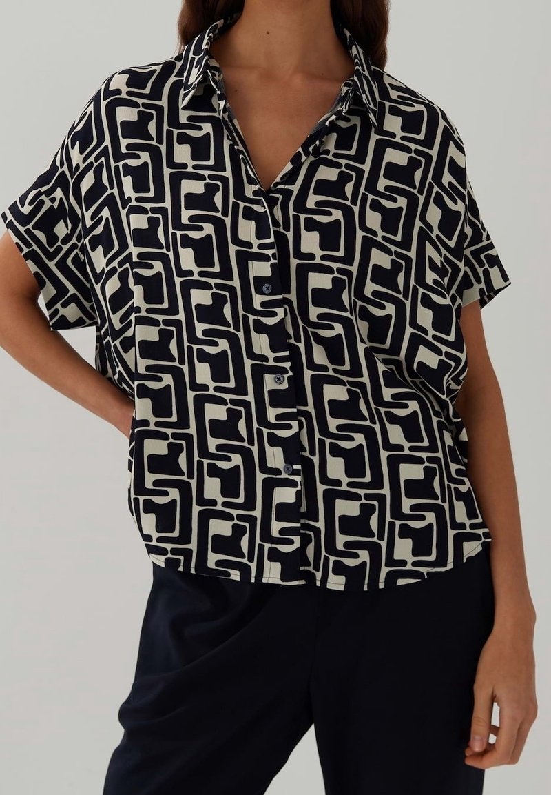 Chemise à manches courtes avec un motif géométrique noir et ivoire, coupe décontractée et col classique. Fabriquée en tissu léger.