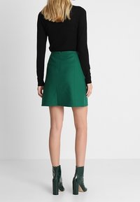 Jupe A-line verte en tissu texturé, avec une fermeture éclair au dos. Associée à un haut noir à manches longues et des bottines à talons vernis vertes.