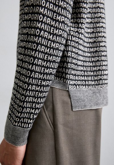 Pulover tricotat alb-negru cu un model repetitiv al logo-ului "Emporio Armani", având un guler și o margine texturată cu rigle.