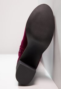 Semelle et talon en caoutchouc noir d'une chaussure en daim bordeaux posée verticalement sur une surface blanche.