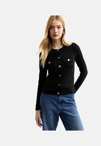 Haut noir à manches longues avec des boutons dorés, doté de deux poches poitrine et d'un col ras du cou, associé à un jean en denim bleu.