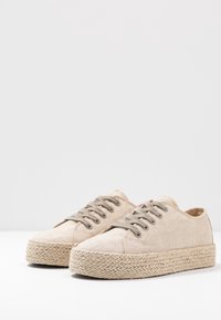 Baskets en toile beige avec semelles compensées tissées épaisses et lacets beige assortis sur un fond clair uni.