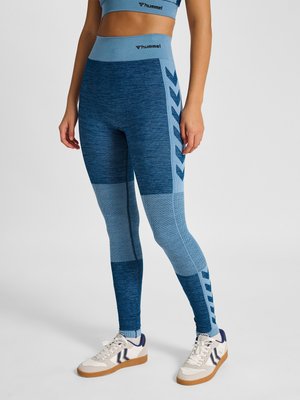Frau trägt blaue nahtlose Leggings mit gemusterten Einsätzen und seitlichen Chevron-Streifen, kombiniert mit weißen Sneakers auf einfarbigem Hintergrund.