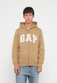 Bežová mikina na zips s bielym logom "GAP" na hrudi, s prednou kapsou a nastaviteľnou kapucňou so šnúrkou. Štandardný strih.
