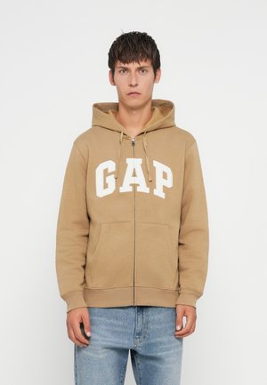 Beige zip-up hoodie with white "GAP" logo on the chest, featuring a front pocket and adjustable drawstring hood. Standard fit.
