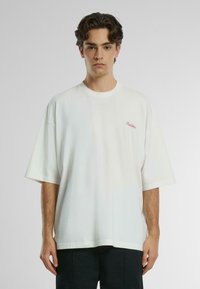 Camiseta de algodón blanca de gran tamaño, con cuello redondo y mangas cortas, que presenta un pequeño logo rojo en la parte izquierda del pecho.