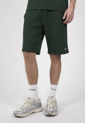 Man draagt donkergroene knielange shorts, witte sportsokken met zwarte tekst en zilveren New Balance hardloopschoenen, staand tegen een effen achtergrond.