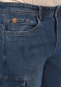 Jeans od denima u tamnoplavoj boji s teksturiranim završetkom. Ima prednju džepu s kožnom oznakom i detaljima šivanja duž struka i džepova.
