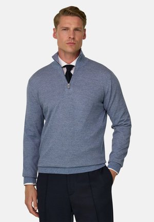 Homme portant un pull bleu à fermeture éclair quart de zip par-dessus une chemise blanche et une cravate noire, avec un pantalon noir, debout devant un fond uni.