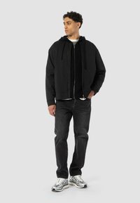 Juodas zip-up megztinis su raišteliais, dėvimas virš šviesiai pilkos crew neck marškinėlių, derinamas su tamsiais džinsais ir metaliniais sportbačiais.