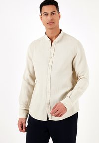 Beige hosszú ujjú gombos ing mandarin gallérral, texturált anyagból és egyenes szegéllyel. Elöl gombokkal és mandzsettás részletekkel rendelkezik.