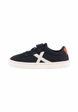 Zapatilla negra de caña baja con suela blanca, detalle lateral blanco en forma de "X", cordones negros y parche naranja en el talón, mostrada en perfil lateral.