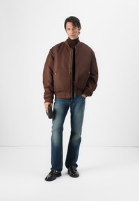 Casaco bomber acolchoado castanho com fecho éclair, bolsos laterais e colarinho canelado, combinado com jeans escuros e sapatos pretos de vestir.