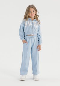 Ljusblått barn tracksuit med en kortärmad zip-up hoodie som har "MALIBU" i vitt, kombinerat med löst sittande byxor, båda i mjukt tyg.