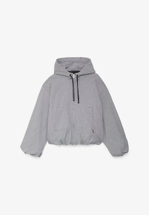 Mujer con chaqueta gris con capucha y cremallera sobre una falda blanca plisada, calcetines blancos y mocasines negros, de pie frente a una pared blanca.