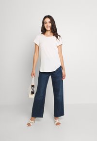 Krømlig kortermet topp, mørkeblå vide jeans, hvit håndveske og lyse høyhælte sko, med solbriller i hånden.