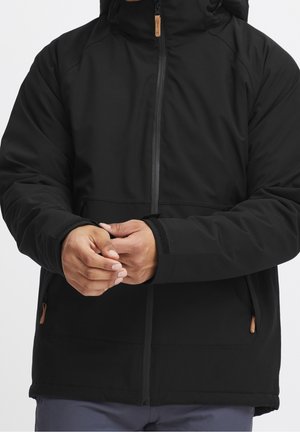 Mann trägt eine schwarze Jacke mit Reißverschluss und Kapuze und befestigt den Klettverschluss am linken Ärmel, die Hände in Taillennähe gehalten.