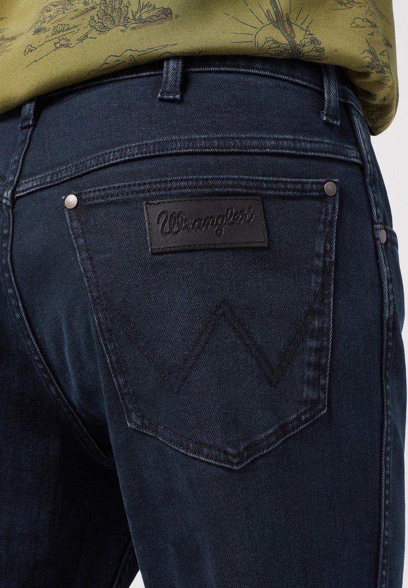 Vaqueros de mezclilla oscura con un diseño distintivo en los bolsillos traseros, con una etiqueta de cuero de Wrangler y costuras contrastantes en los bolsillos.