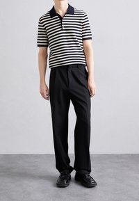 Muž oblečený v černo-bílém pruhovaném polokošili s krátkým rukávem, širokých černých kalhotách a černých kožených mokasínech stojí na šedé podlaze.