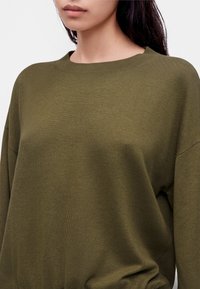 Jeune femme portant un pull en maille vert olive à col rond, visible du cou jusqu'au milieu du torse sur un fond uni.