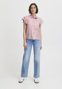 Blusa rosa chiaro con dettaglio a fiocco al colletto, maniche a sbuffo corte, abbinata a jeans a gamba larga blu chiaro e sneakers bianche.