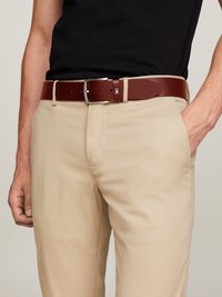 Ceinture en cuir brun avec une boucle en métal argenté, portée sur un pantalon beige. La ceinture présente une forme rectangulaire et un design minimaliste.